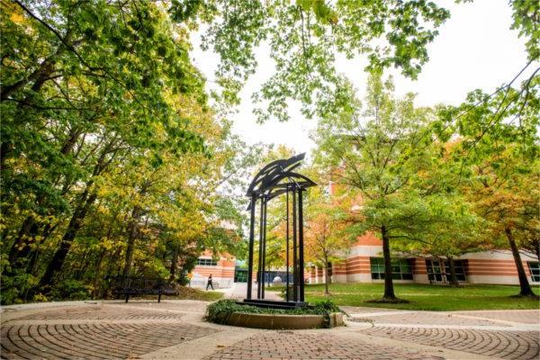 A sculpture sits on a brick walkway, surrounded by trees.